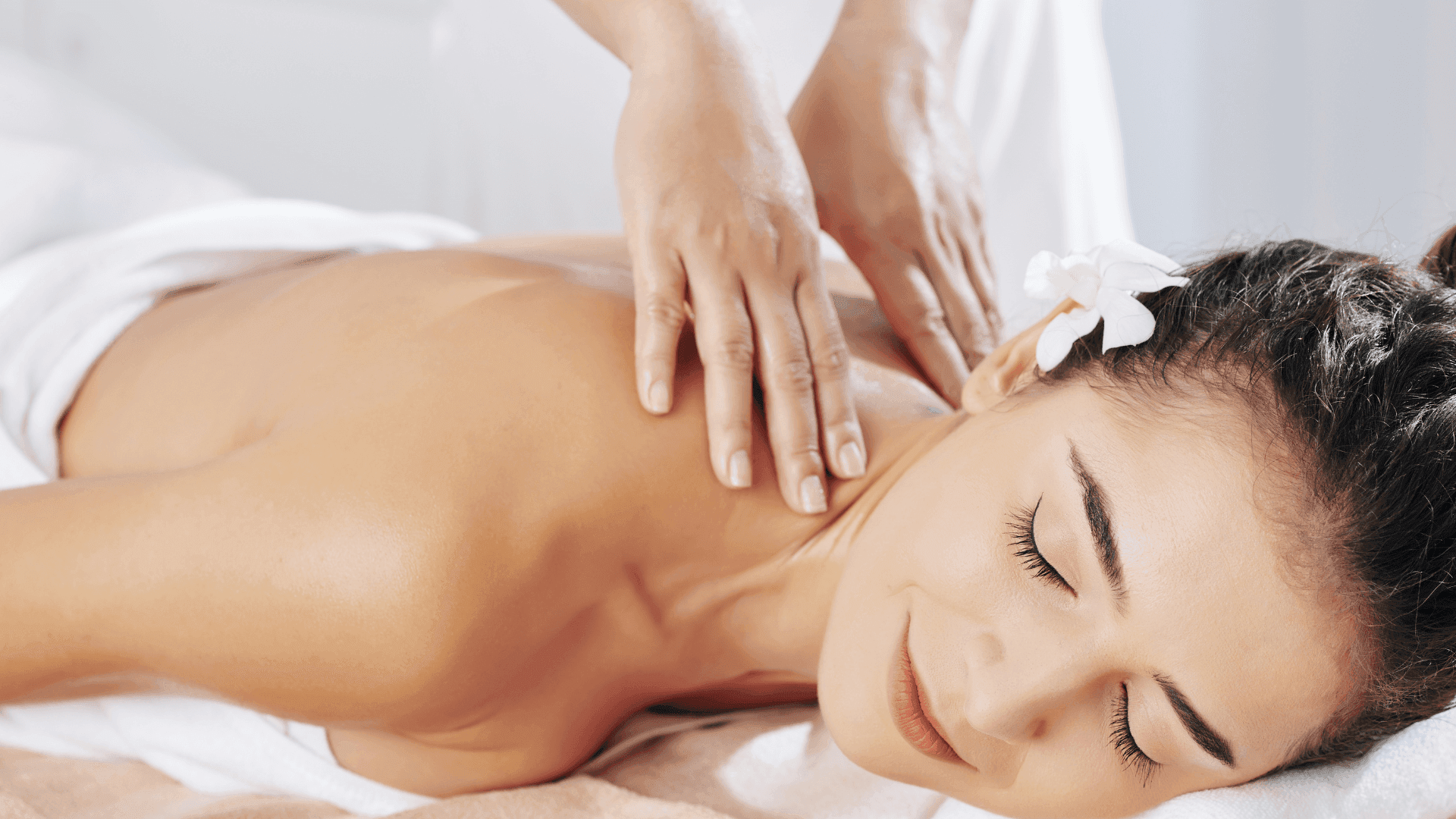 Estetista che massaggia la schiena di una donna sdraiata sul lettino, a occhi chiusi, con un fiore bianco tra i capelli