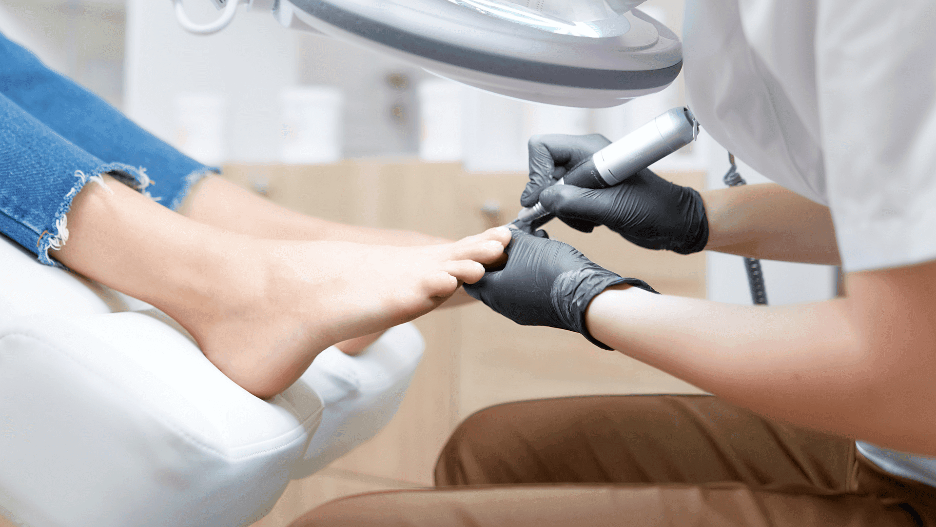 Pedicure: operatrice con guanti neri lavora le unghie del piede di una cliente con una fresa elettrica, sotto la lampada