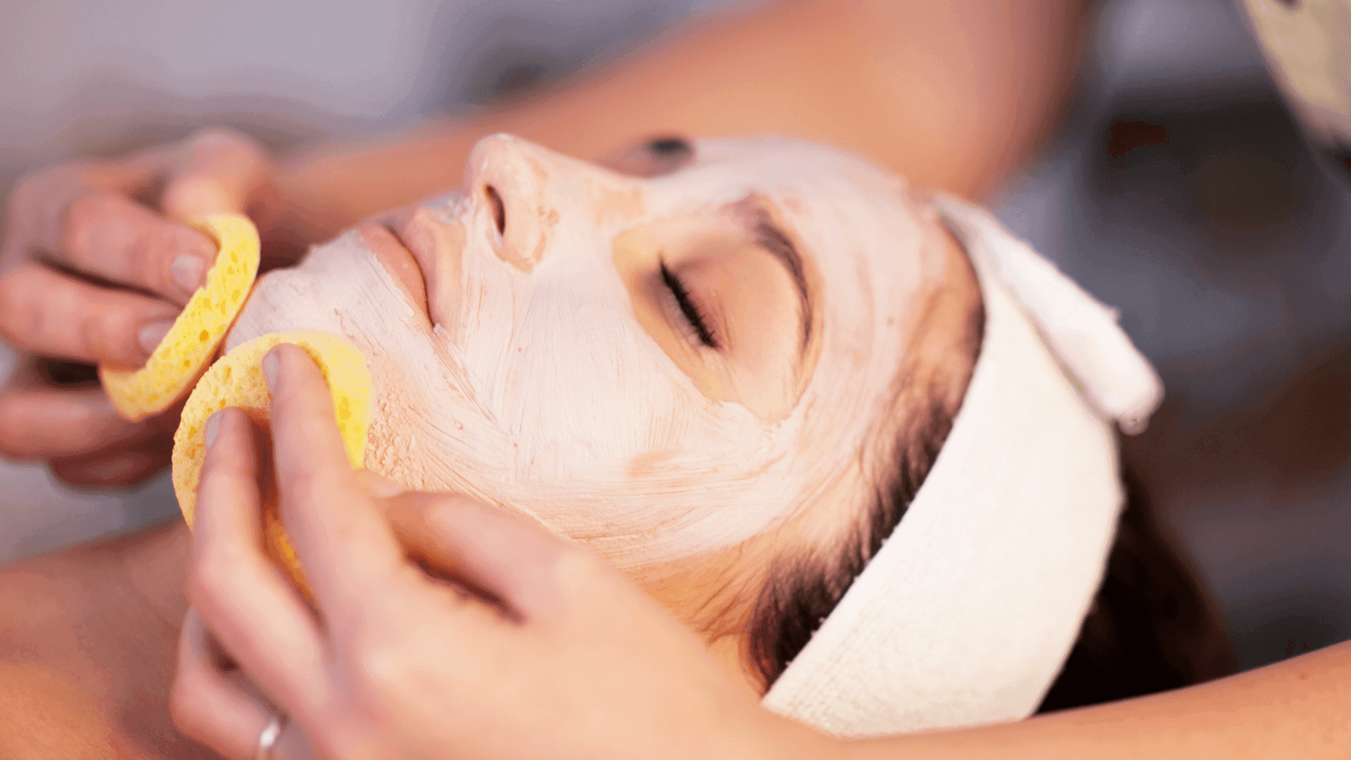 Pulizia del viso: l’operatrice rimuove la maschera con due spugnette dal volto di una cliente con fascia bianca tra i capelli