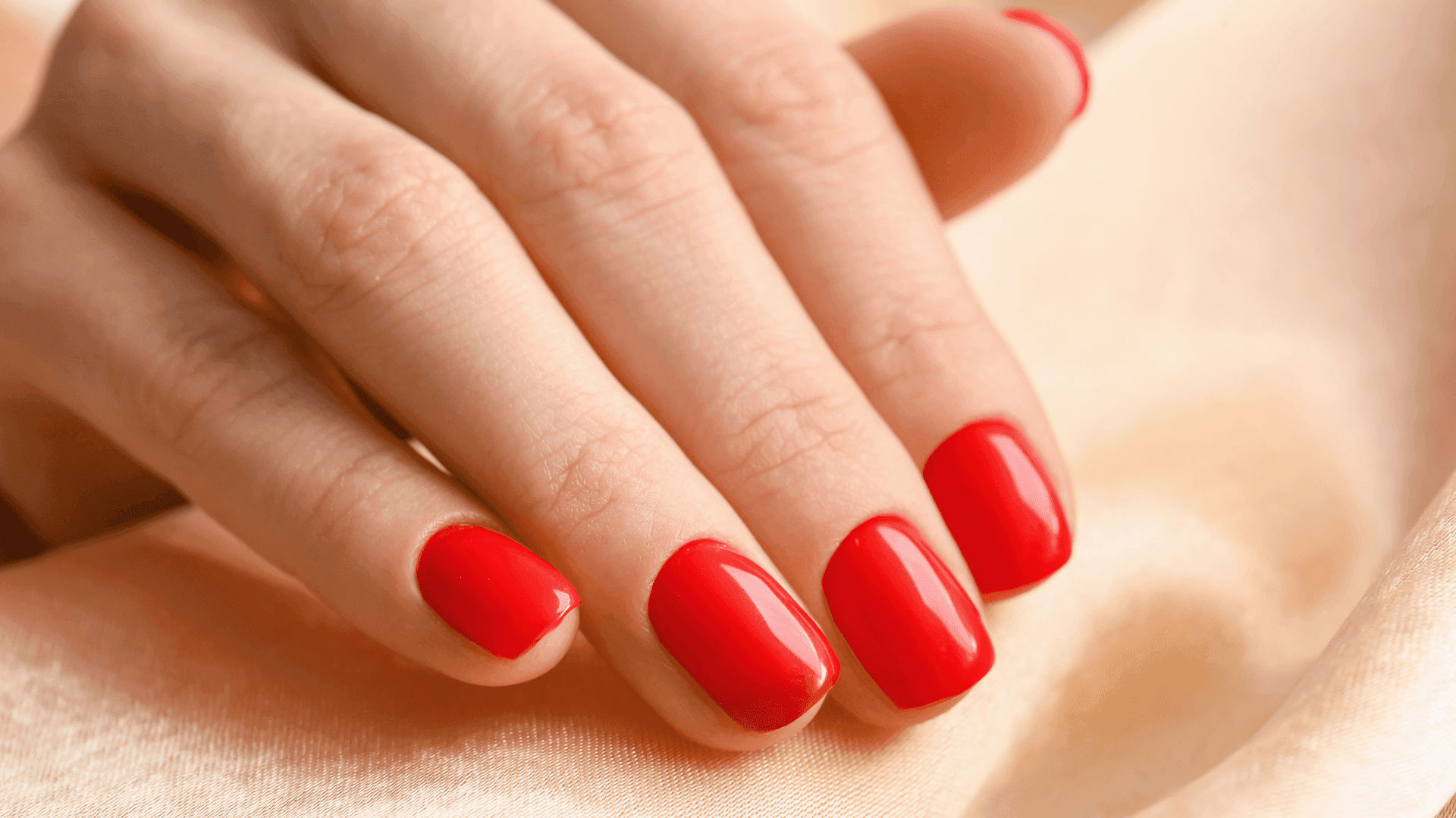 Unghie corte smaltate di rosso brillante con smalto semipermanente, mano appoggiata su un tessuto color crema