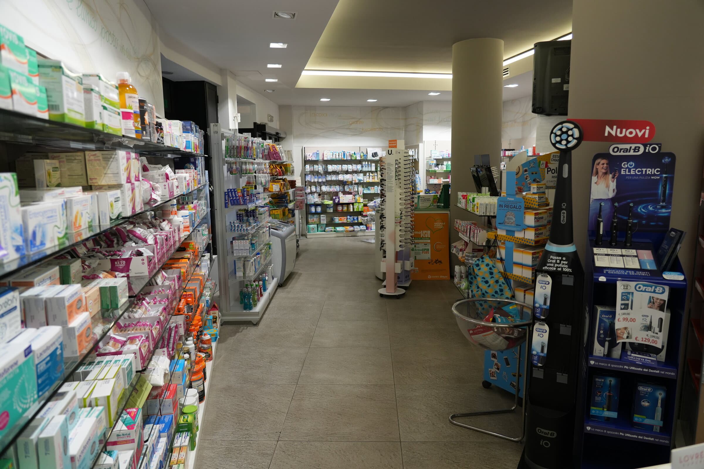 Corsia interna della Farmacia Dova tra gli scaffali di prodotti per la cura della persona, con un espositore di spazzolini elettrici sulla destra